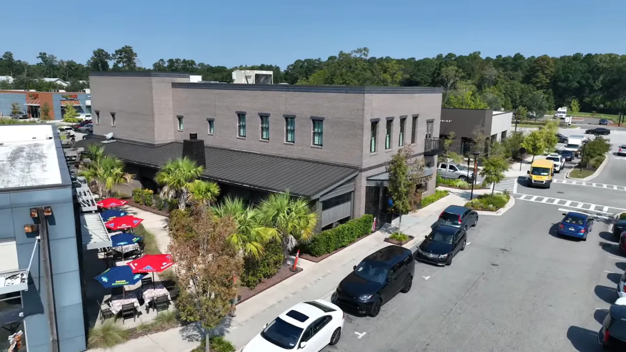 Aerial/drone view of a neighborhood street with shops, outdoor seating, trees and parked cars