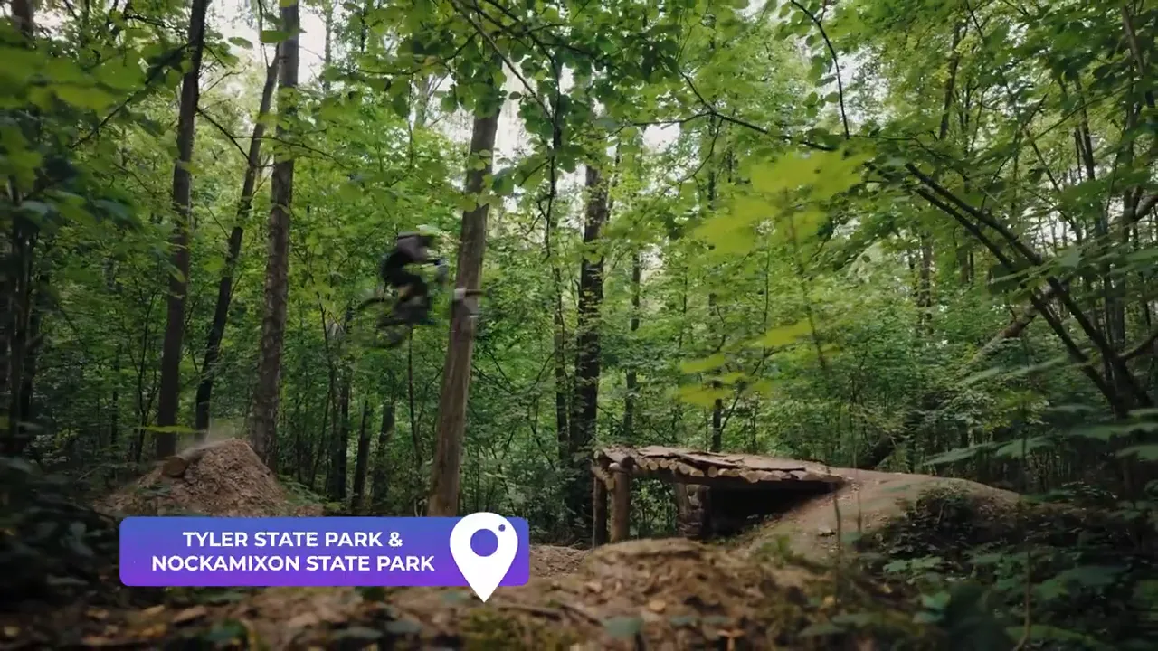 Forest trail with wooden ramp and an on-screen label reading 'TYLER STATE PARK & NOCKAMIXON STATE PARK', a biker mid-air in the background