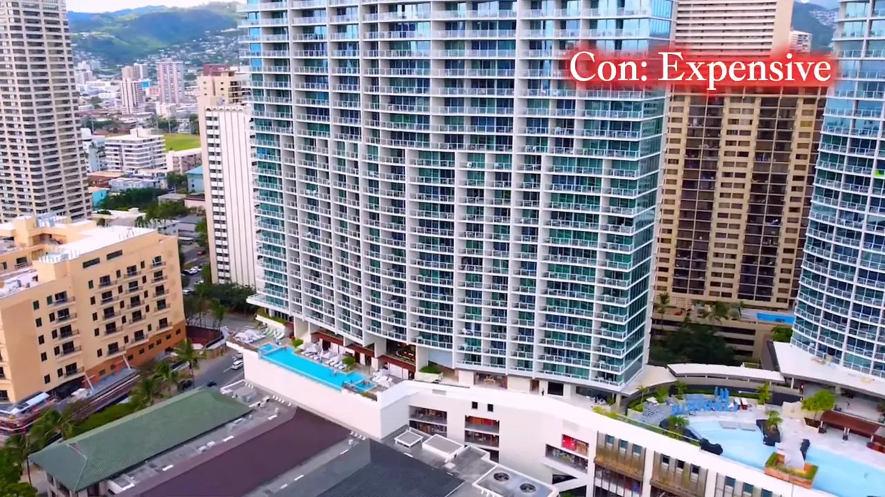 Aerial view of tall condominium towers in Waikiki with pool decks and the text overlay 'Con: Expensive'