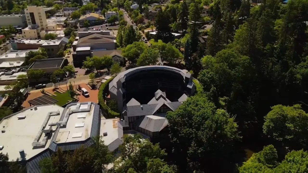 High-quality aerial photo showing the Oregon Shakespeare Festival outdoor amphitheater surrounded by trees and downtown buildings.