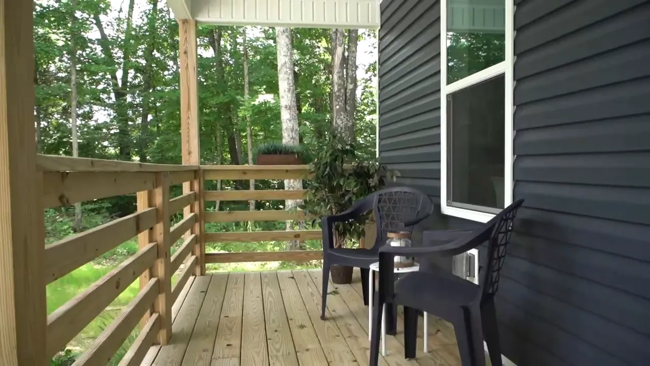 Covered front porch with wood decking, railing, and two black chairs beside the exterior wall