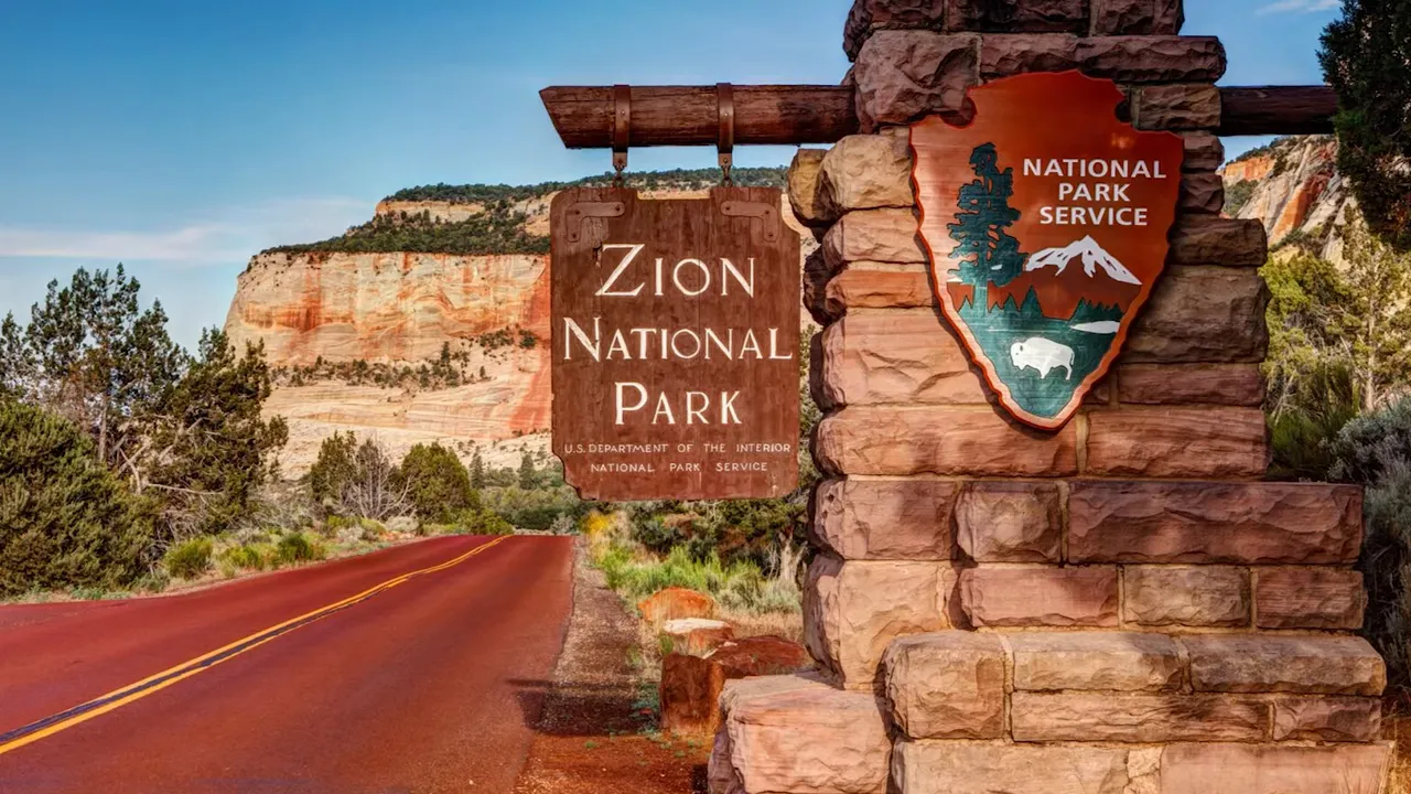 Zion National Park entrance sign beside a paved road and red rock landscape