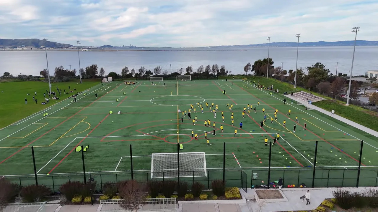 Aerial view of multiple youth soccer drills and games on waterfront sports fields with goals and players in bright jerseys
