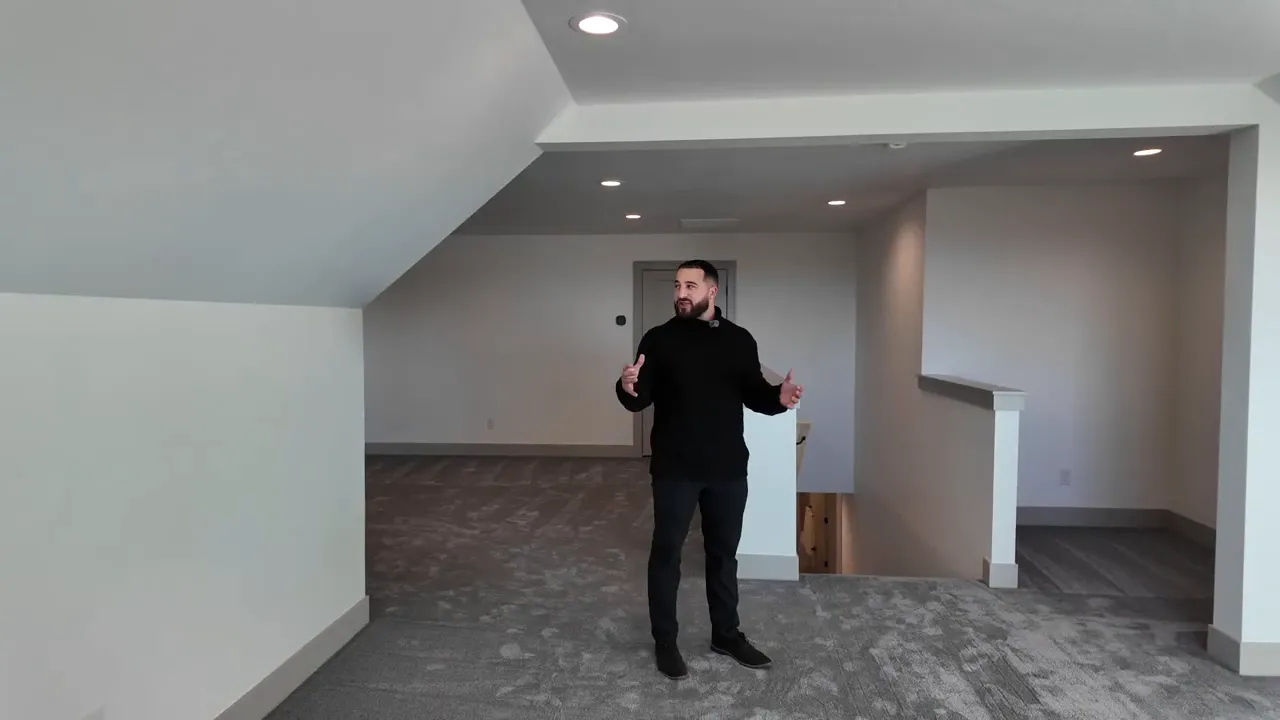 Open upstairs bonus room with sloped ceiling, recessed lighting, carpeted floor and presenter gesturing near the stairwell