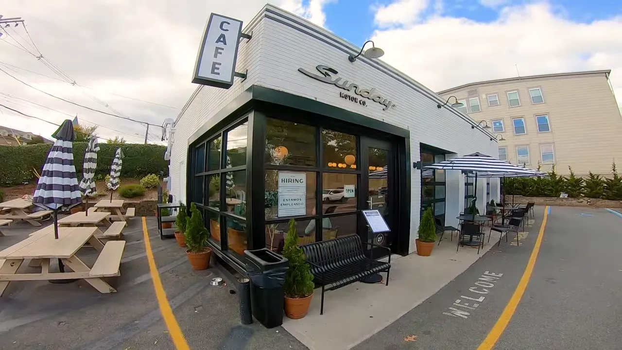 Exterior of a small downtown café with picnic benches, umbrellas, potted plants and a 'CAFE' sign.