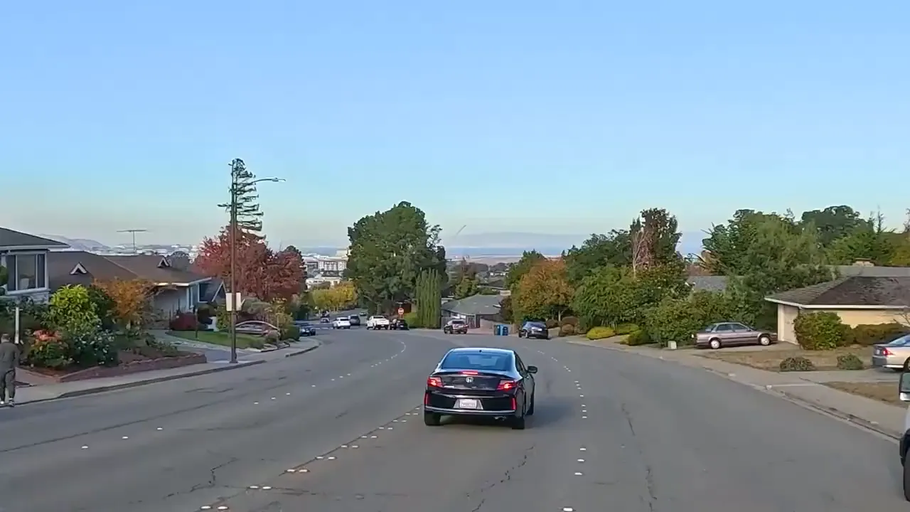 Street-level view down a sloping residential street with parked cars and distant bay and neighborhood views