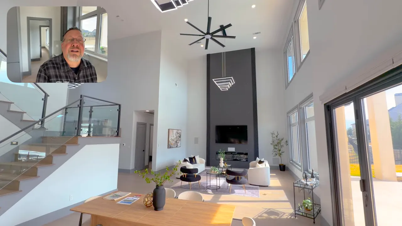 Wide-angle view of the two-story living room showing the tall media/fireplace wall, high ceilings, seating area, dining table and sliding doors to the patio; video host inset top-left.