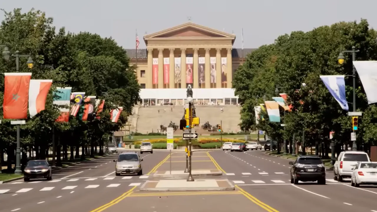 View down Ben Franklin Parkway toward the Philadelphia Museum of Art with flags lining the avenue and cars on the road.