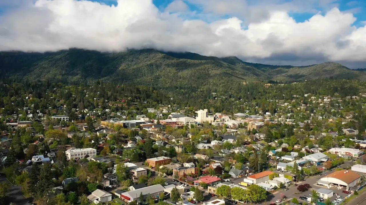 Aerial panorama of Ashland, Oregon — downtown and residential areas with tree cover and forested hills beneath a partly cloudy sky.