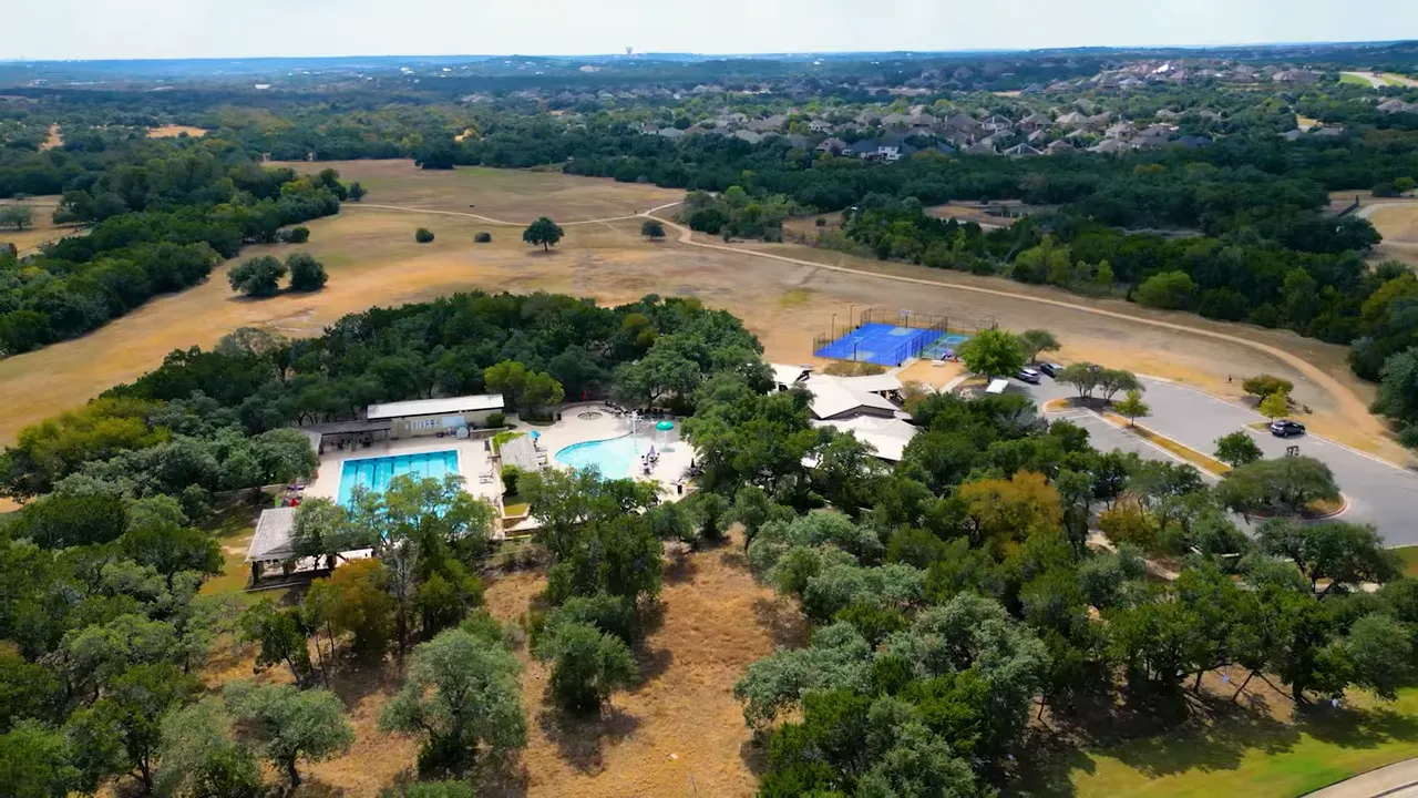 Aerial shot of a community center with two swimming pools, tennis courts and parking surrounded by trees and open fields