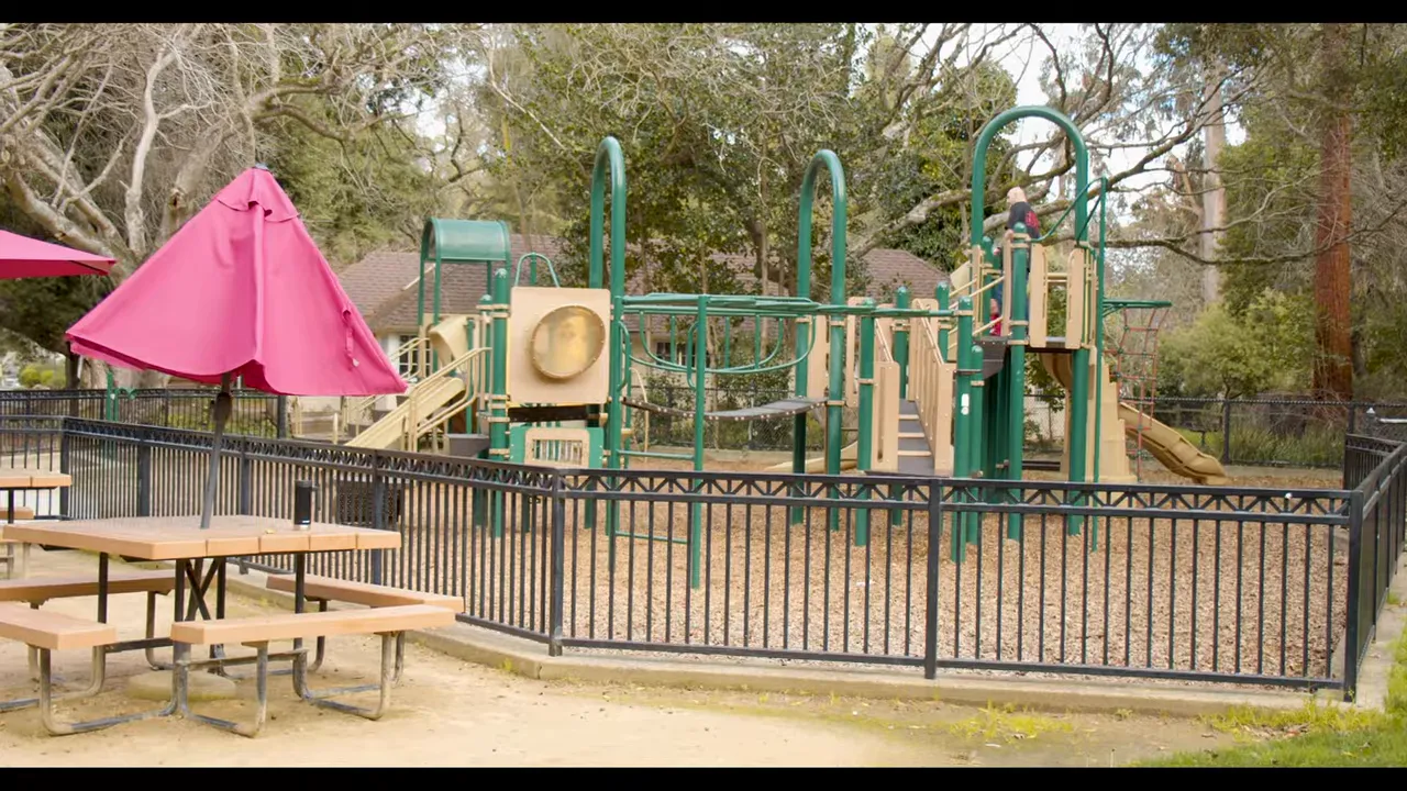 playground behind a fence with picnic tables and pink umbrellas