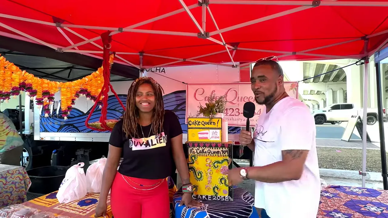 Vendor and interviewer under a red market tent at a Jacksonville arts market with colorful display and signage