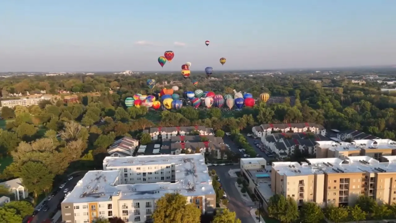 Wide aerial shot of dozens of colorful hot air balloons over a tree-lined park and nearby apartment buildings