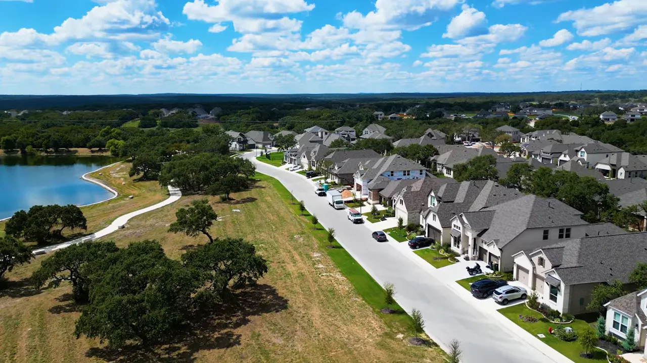 Aerial view of a suburban street running alongside a lakeside walking path with single-family homes and oak trees in an Austin-area neighborhood.