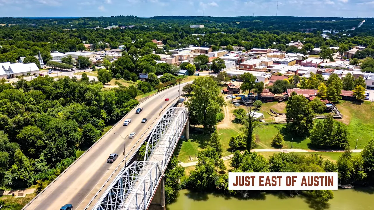 High-angle aerial of a bridge crossing a river with a small town and green tree canopy just east of Austin; overlaid text reads 'JUST EAST OF AUSTIN'.