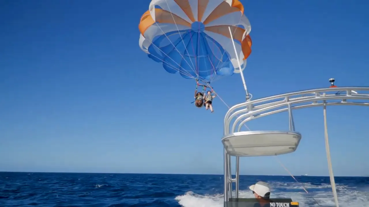 two people parasailing beneath a blue and orange canopy over deep blue ocean, photographed from the boat