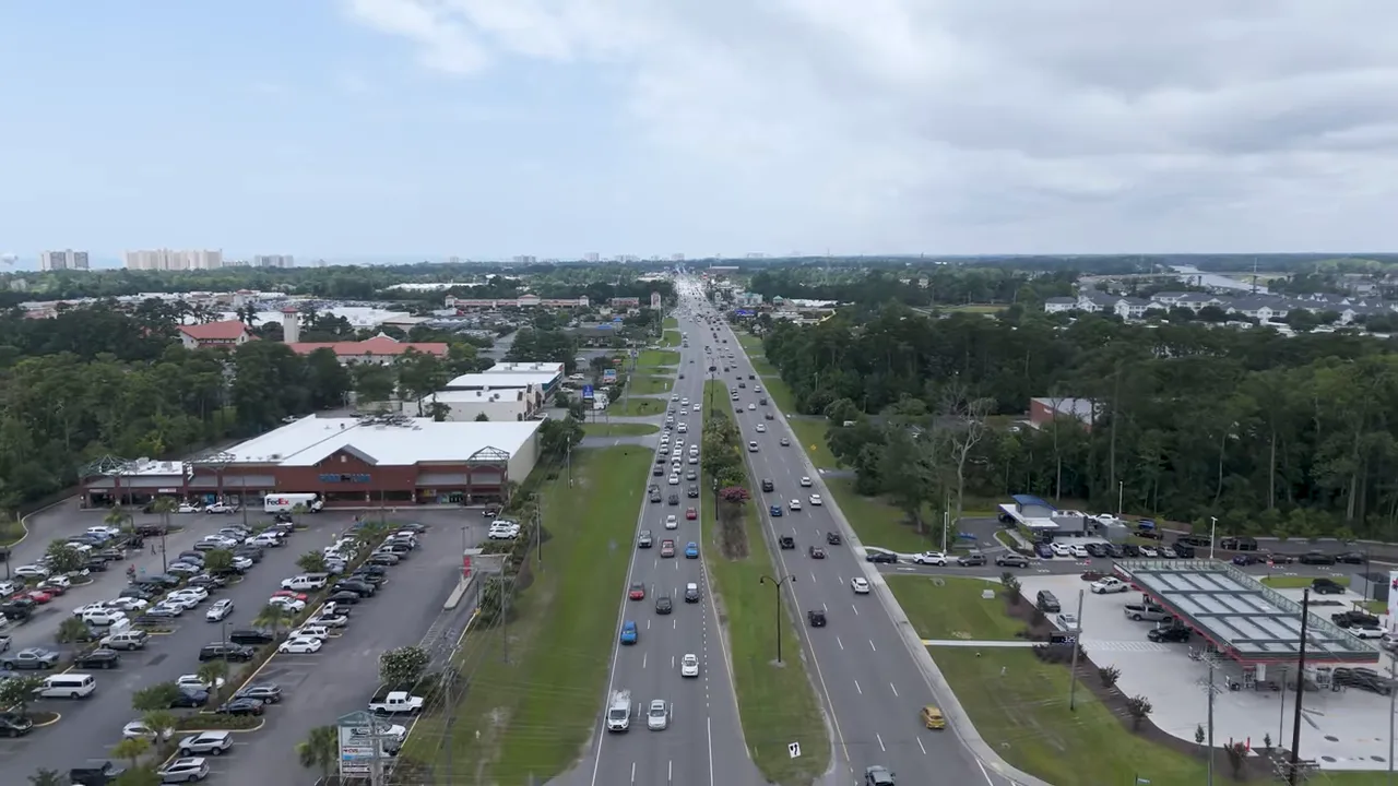 Aerial view of a multi-lane highway with traffic, adjacent shopping centers and green median in a coastal metro area.