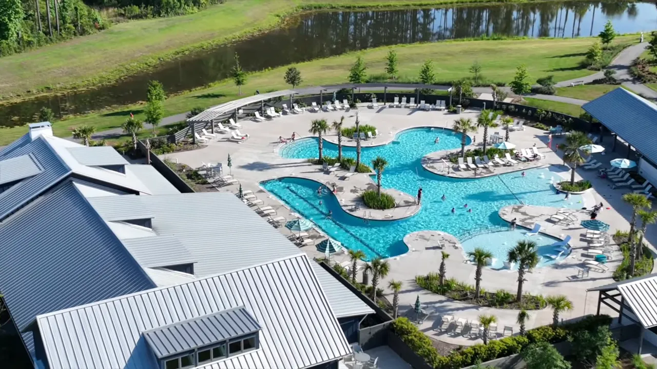 Aerial view of a resort-style community pool and clubhouse with lounge areas, palm trees and a nearby pond