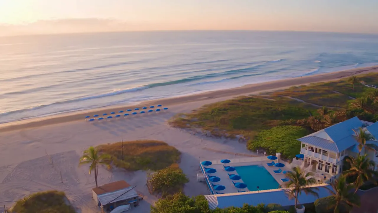 Aerial sunrise view of Delray Beach showing rows of blue beach umbrellas, ocean waves, and a beachfront pool and club