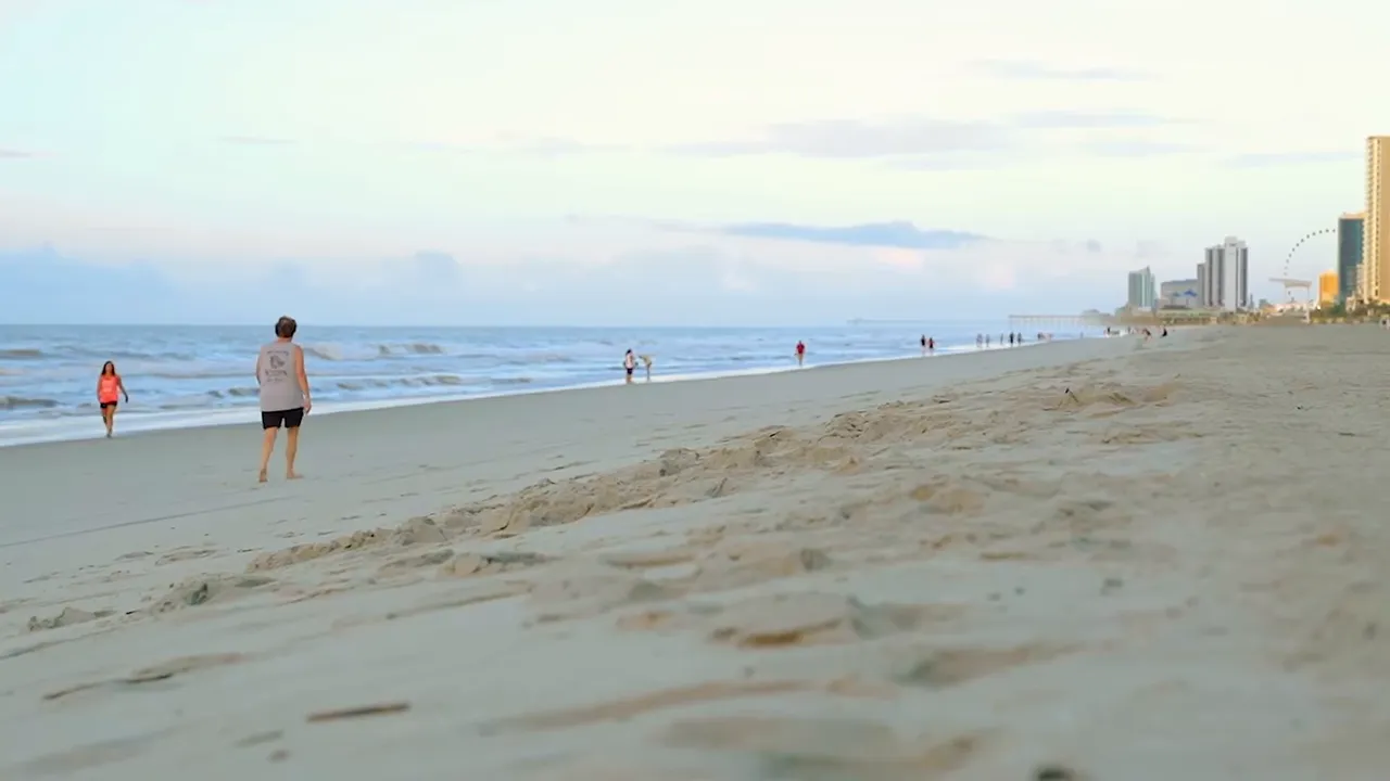 Wide sandy Myrtle Beach shoreline with people walking along the water and high-rise hotels visible on the horizon.