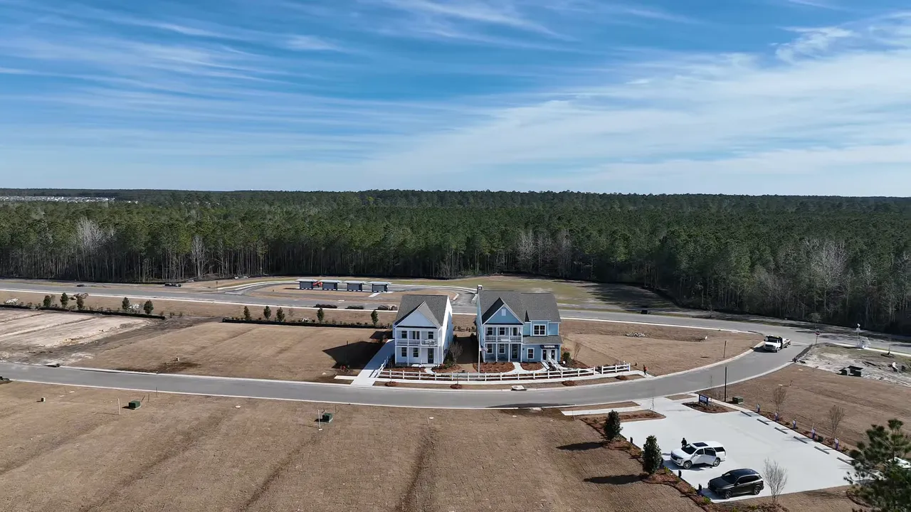 Wide aerial/drone view of two houses on a curved road with large yards, adjacent empty lots and a tree line — illustrating neighborhood layout and maintenance.