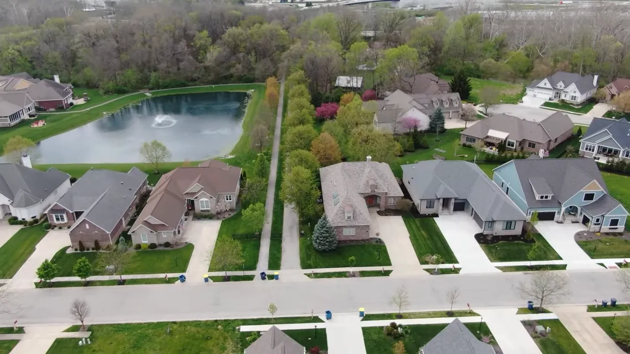 Aerial shot of an Indianapolis-area suburban neighborhood showing homes, a pond with fountain, green lawns and a walking path between properties.