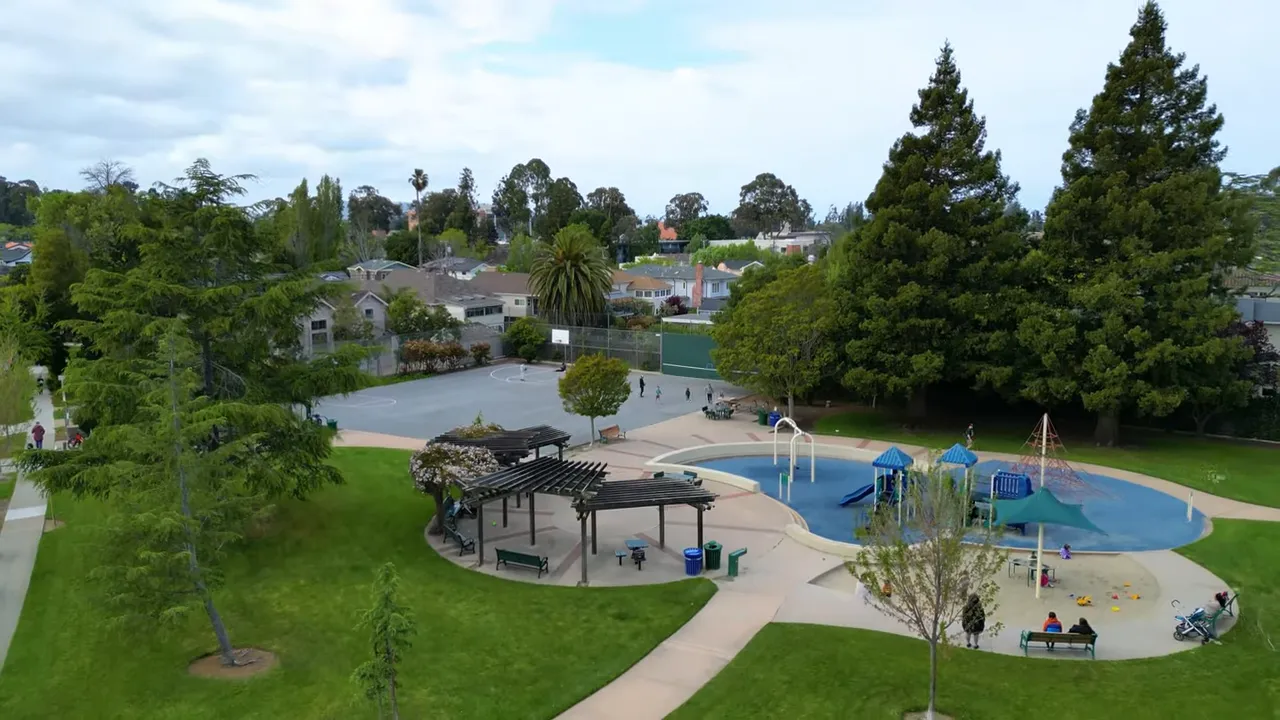 Community park with playground, splash area, basketball court and large trees, viewed from above