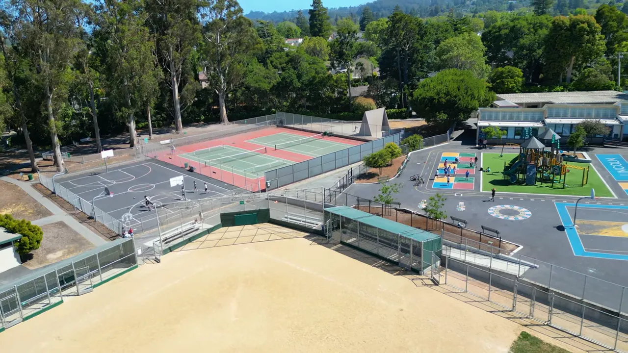 Aerial view showing a baseball field, fenced basketball courts, tennis courts and a colorful playground at a school campus.
