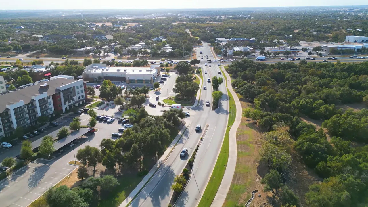 Aerial shot of a multi-lane road and nearby commercial buildings with a large preserved wooded greenbelt and a sidewalk/trail.