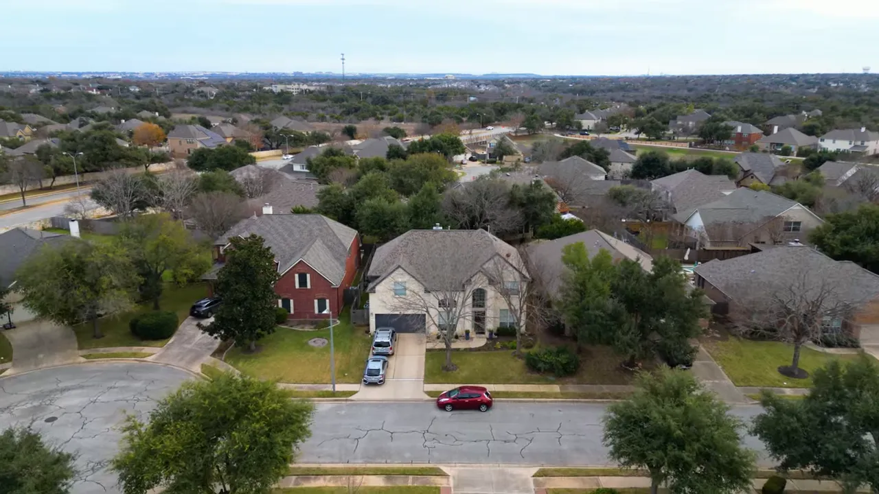 Aerial drone view of a suburban neighborhood with single-family houses, yards, trees, and a main road in the distance