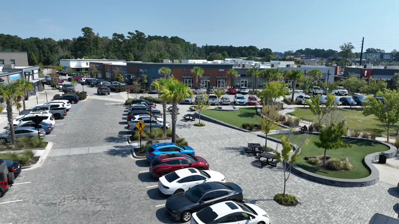 Mixed-use town center plaza with shops, parked cars, palm trees, landscaped green areas, and benches viewed from above
