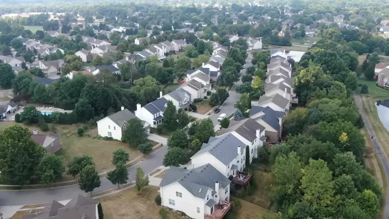 High-angle aerial of a suburban residential street with rows of single-family homes, backyards, trees and nearby pond.