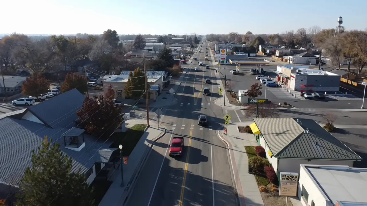 Aerial view of a small town main street with cars, storefronts, a Subway sign, a Shell station and a distant water tower.