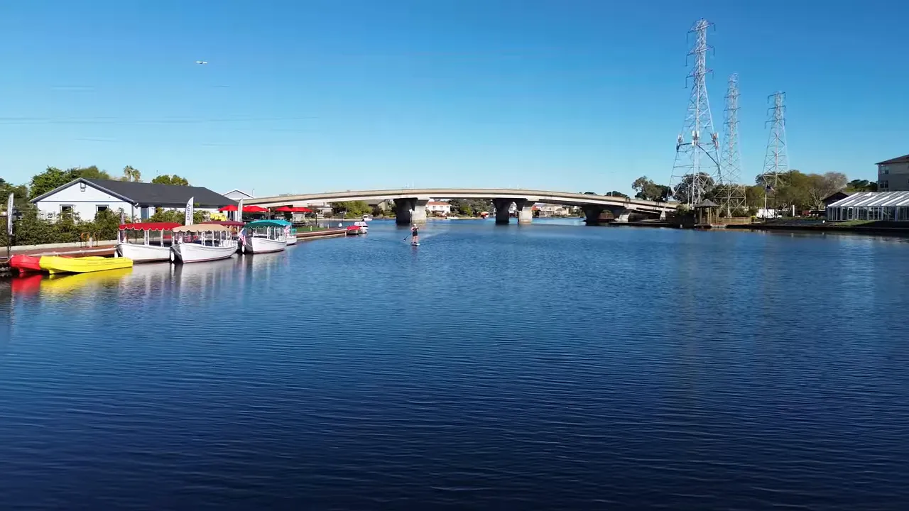 Paddleboarder and docked small boats on a calm blue lagoon with a bridge in the background