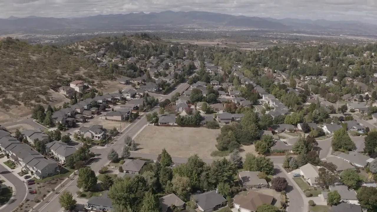 Wide drone panorama of Southern Oregon neighborhoods, open lots and surrounding hills giving regional context.