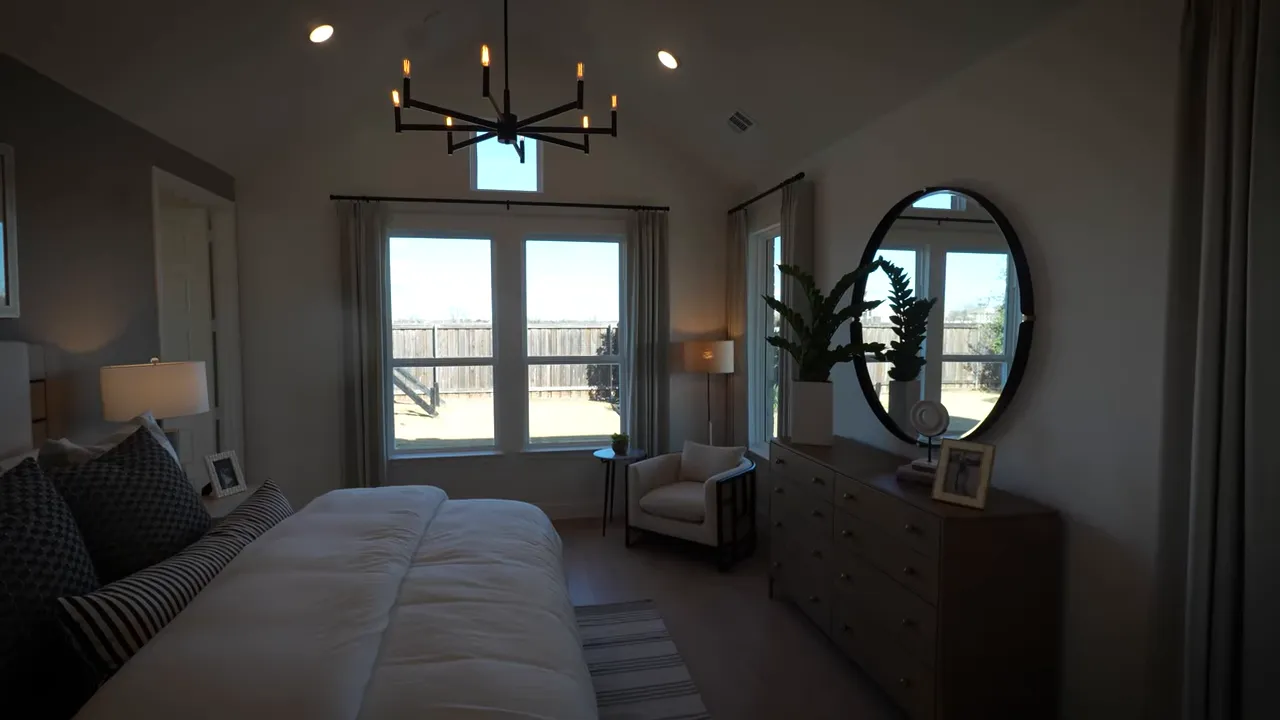 Wide view of primary bedroom with vaulted ceiling, chandelier, bed, large windows and dresser with round mirror