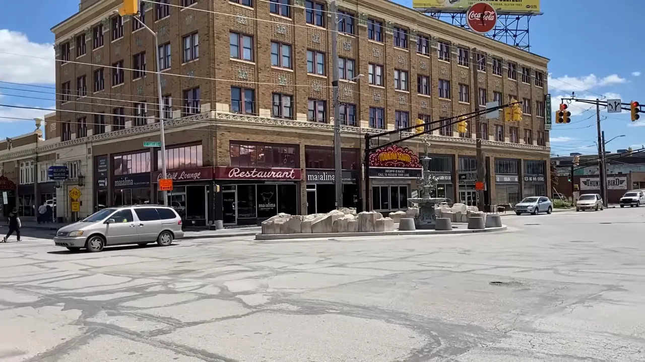 Historic mixed-use building and small plaza at a walkable Indianapolis intersection, showing streetscape, fountain, and traffic.
