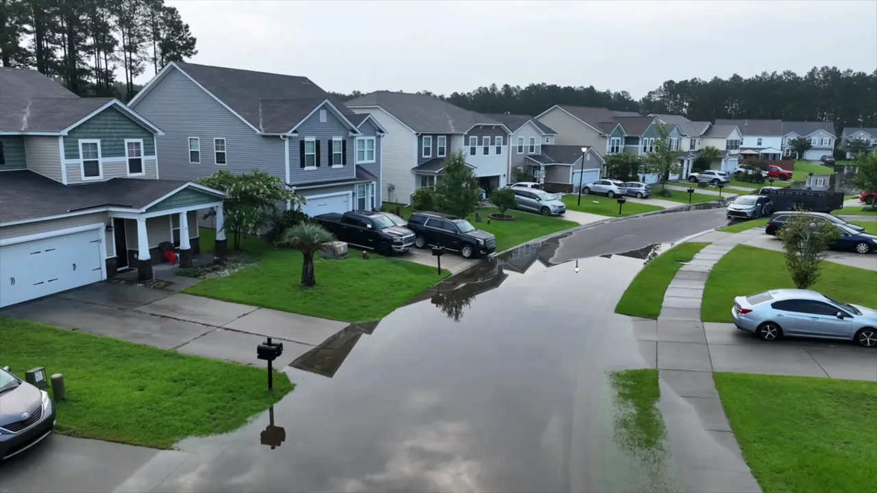 High‑resolution aerial photo of a residential street with extensive standing water reflecting houses and sky, showing drainage issues.