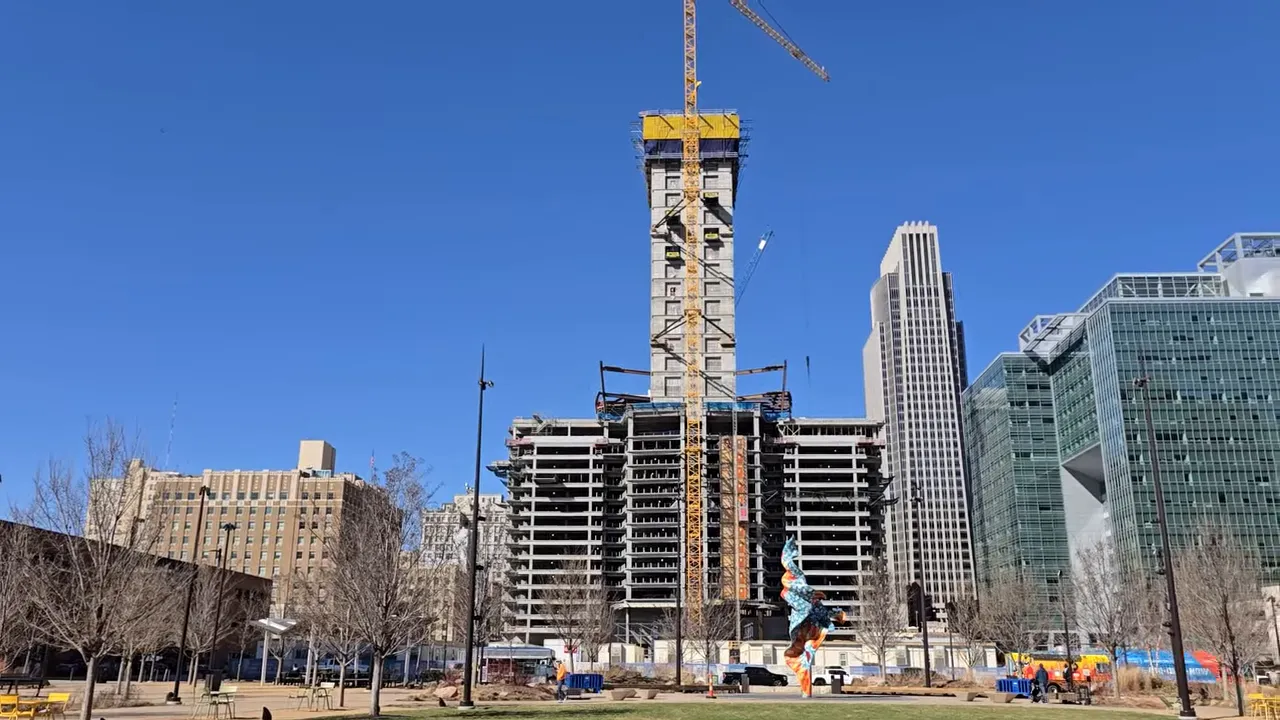 Downtown Omaha high-rise tower under construction with cranes and surrounding buildings under a clear blue sky.