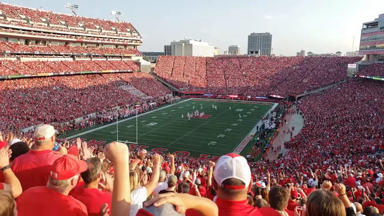 Packed college football stadium filled with fans wearing red watching a daytime Nebraska Huskers game.