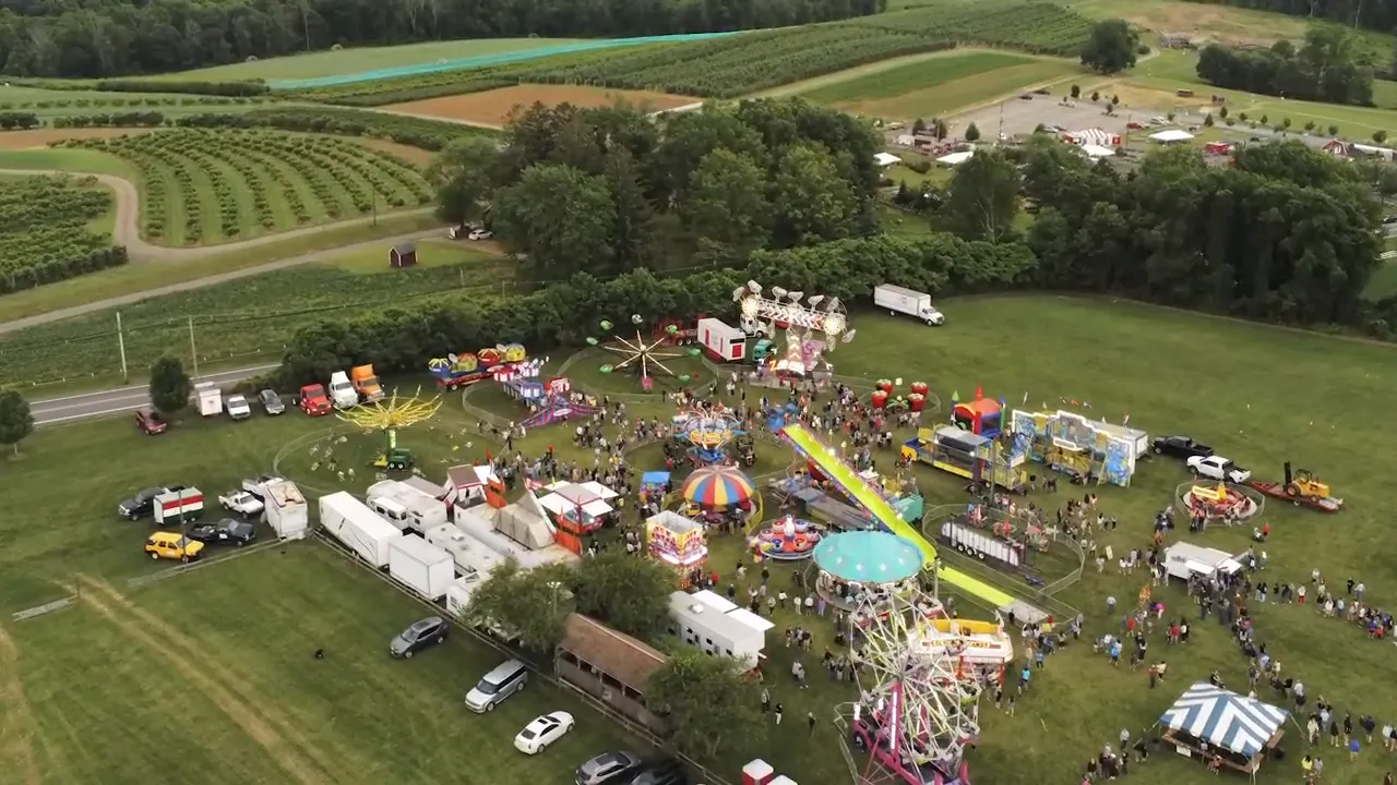 High-resolution aerial view of a carnival with ferris wheel, colorful rides, tents and orchard fields in the background