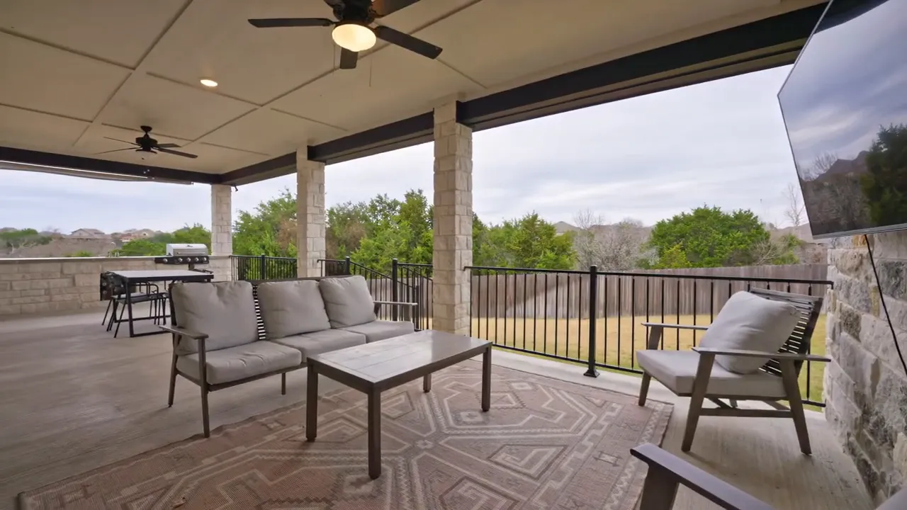 Covered outdoor living area with sofa, dining table, ceiling fans, mounted TV and a grill overlooking the backyard.