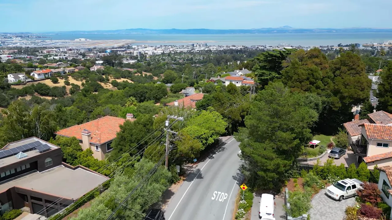 Wide aerial view over Burlingame Hills showing spacious lots, tree canopy, homes and the bay on the horizon.