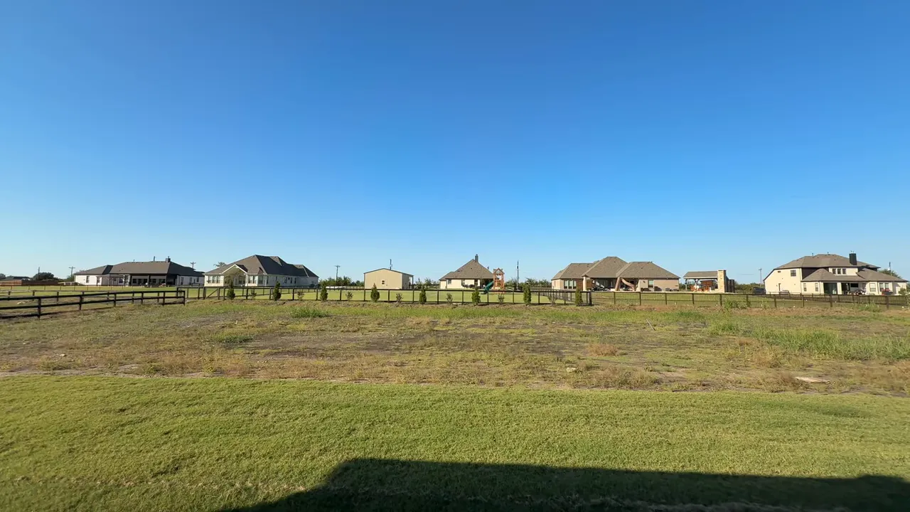 Clear wide-angle view of an acre lot with maintained grass strip in front, open yard area and houses along the fence under a blue sky