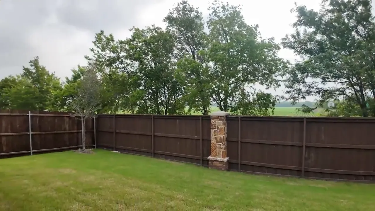 Fenced grass backyard with stone fence columns trees and open land beyond