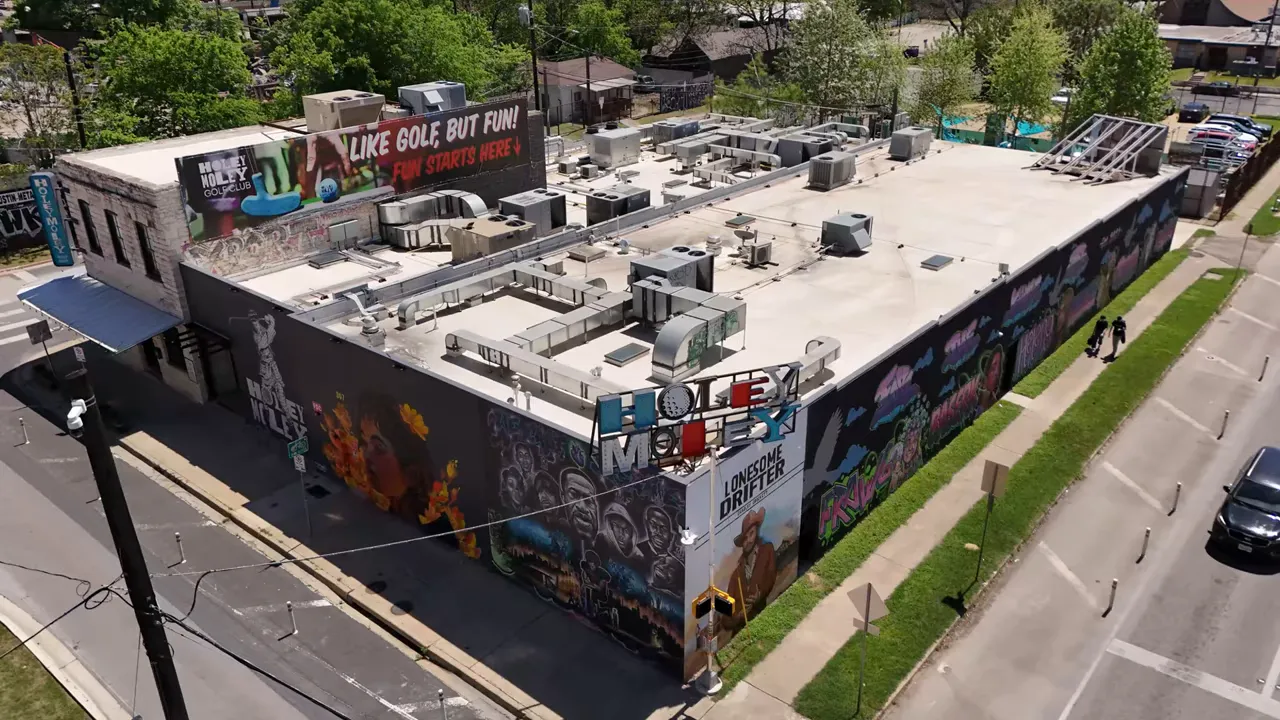 Aerial/angled view of the Holey Moley mini golf building covered in colorful murals with a rooftop billboard reading 'LIKE GOLF BUT FUN!'