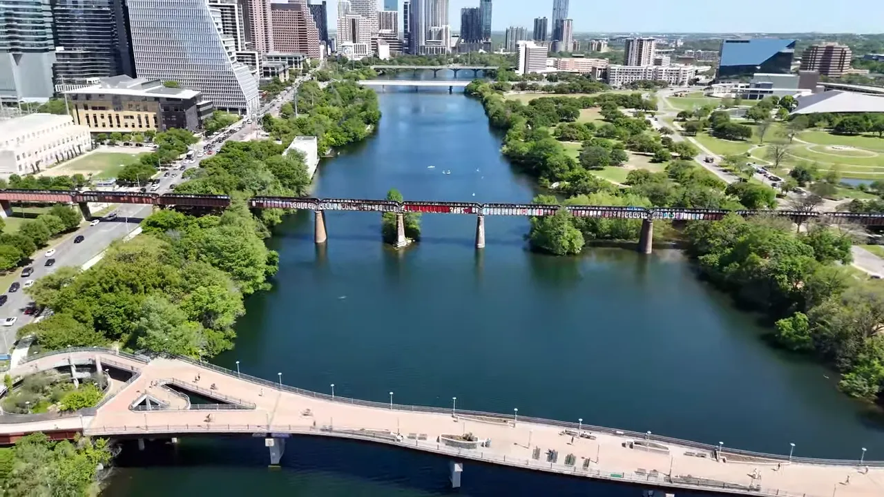 Aerial view showing the Pfluger pedestrian bridge and the graffiti-covered rail bridge crossing Lady Bird Lake with downtown Austin in the distance