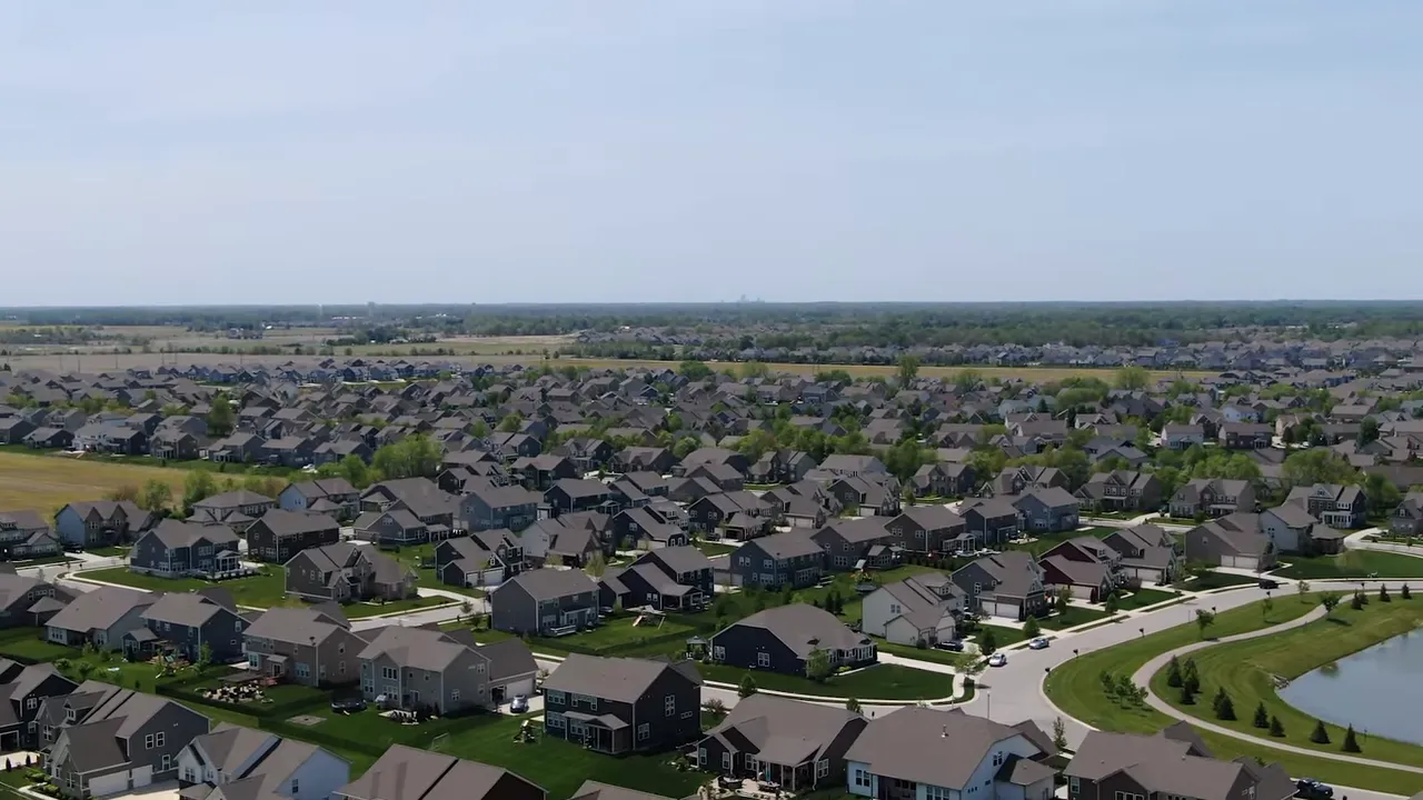 Wide aerial view of Indianapolis‑area suburbs showing many houses, curving streets, green yards and a neighborhood pond.