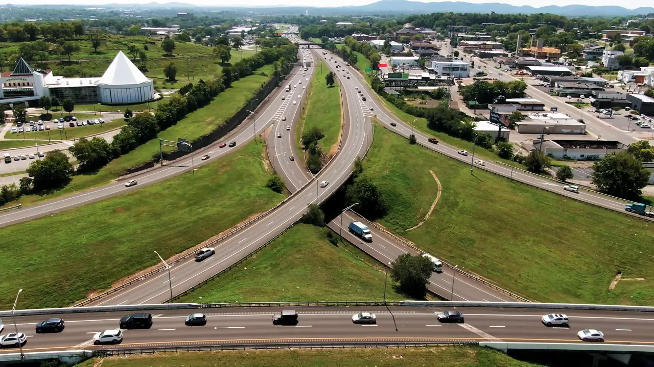 Daytime aerial view of a multi-lane highway interchange, surrounding commercial areas, and green fields.
