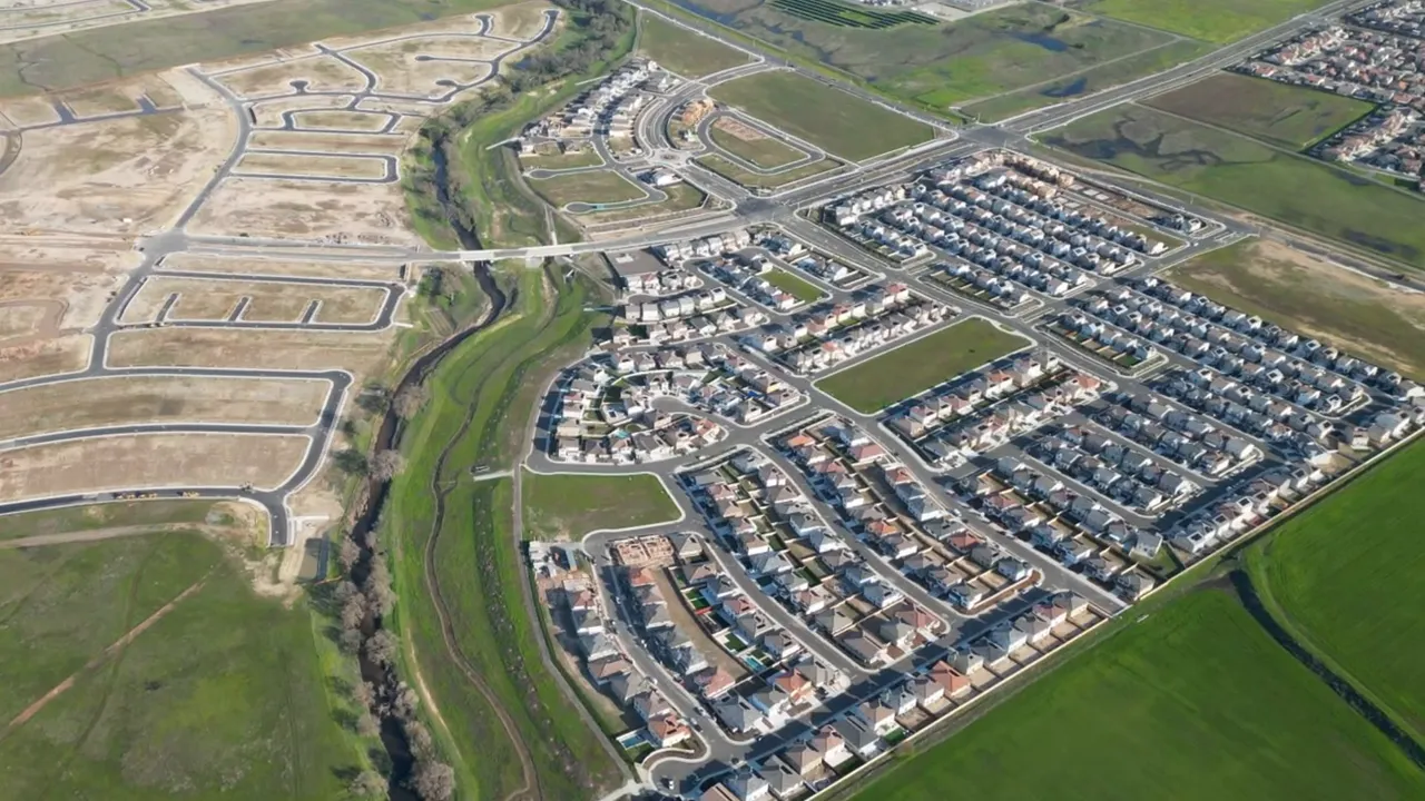Daytime drone view showing built homes, streets, and adjacent undeveloped lots in a new housing development.